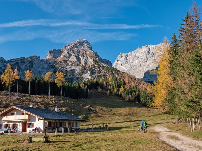 Blick auf einen Wanderweg und eine Hütte mit Menschen davor und Bergen im Hintergrund.