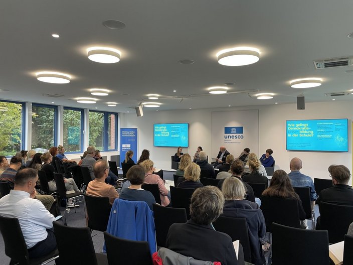 Blick in einen Konferenzraum mit gefüllten Stuhlreihen. Vorne an der Stirnseite diskutieren drei Menschen.