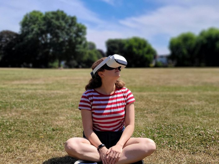 A woman wearing VR glasses sits in a meadow in front of a forest.