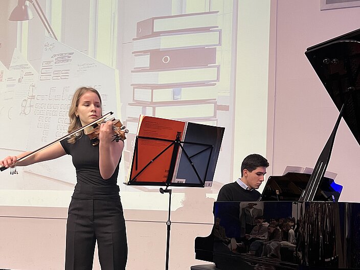 Eine Schülerin spielt die Violine, ein Schüler das Piano auf der Bühne der GSS Tübingen. Im Hintergrund hängt das Plakat der Ausstellung.