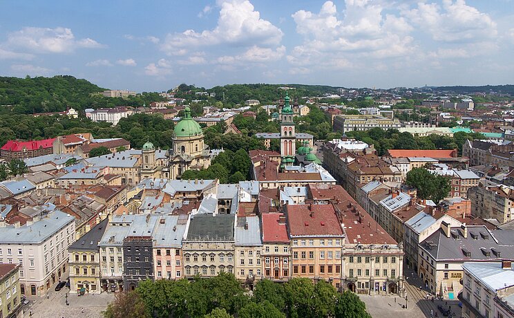 Blick von oben auf die Stadt Lwiw. Man sieht viele alte, sch&ouml;ne H&auml;user, zentral ein sakrales Kuppelgeb&auml;ude und im Hintergrund gr&uuml;ne H&uuml;gel.