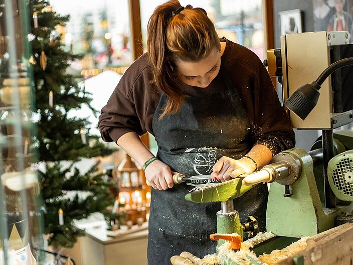 Eine Frau steht in der weihnachtlich geschm&uuml;ckten Schauwerkstatt und arbeitet an einem Holz-Werkst&uuml;ck.