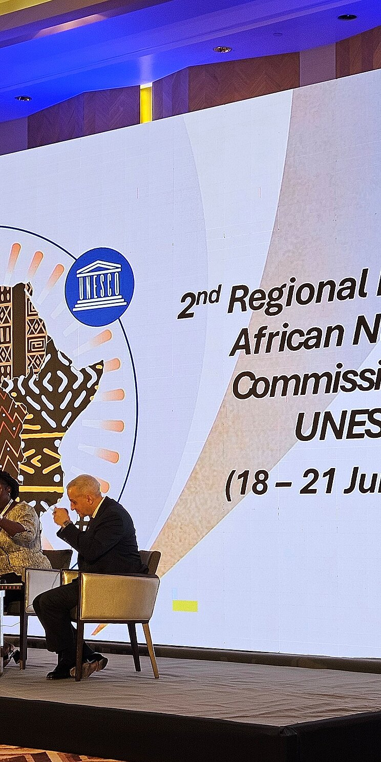 Three people sit on a stage and speak in a panel. Behind them is a large screen with the title ‘Second Regional Meeting of African National Commissions’