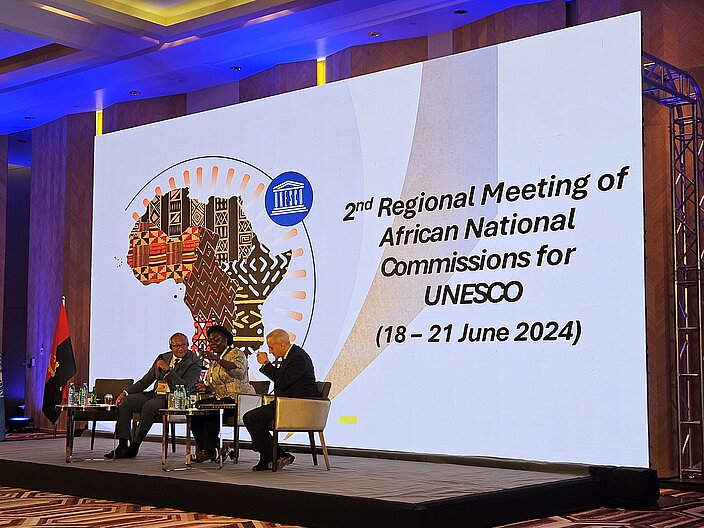 Three people sit on a stage and speak in a panel. Behind them is a large screen with the title ‘Second Regional Meeting of African National Commissions’