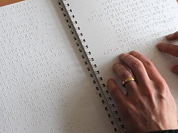 Zwei H&auml;nde ertasten die erhabenen Punkte der Brailleschrift in einem aufgeschlagenen, spiralgebundenen Buch.
