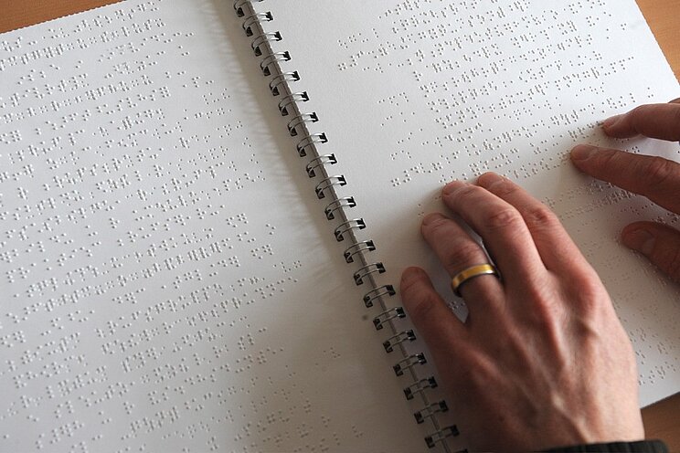 Braille_DBSV Zwei Hände ertasten die erhabenen Punkte der Brailleschrift in einem aufgeschlagenen, spiralgebundenen Buch.