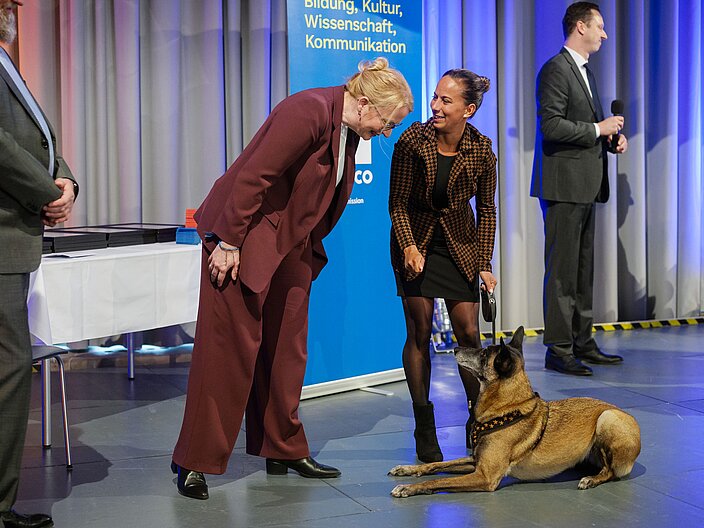 Urkundenverleihung_25_Gebrauchshundewesen Kulturministerin Streichert-Clivot blickt auf einen Hund hinab, der an einer Leine geführt wird.