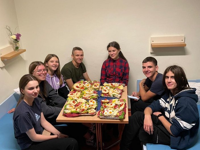 Recreation-Projekt 2023 in Wismar - Willkommenssnack Die Gruppe sitzt um einen Tisch herum auf dem Schnittchen stehen.