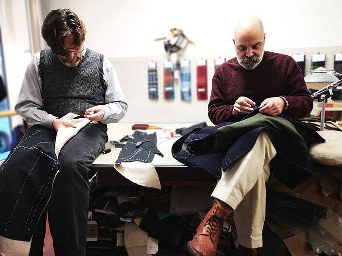 Zwei Schneider sitzen auf einem Tisch. Beide haben Stoffst&uuml;cke in der Hand und arbeiten mit ihren H&auml;nden an den St&uuml;cken
