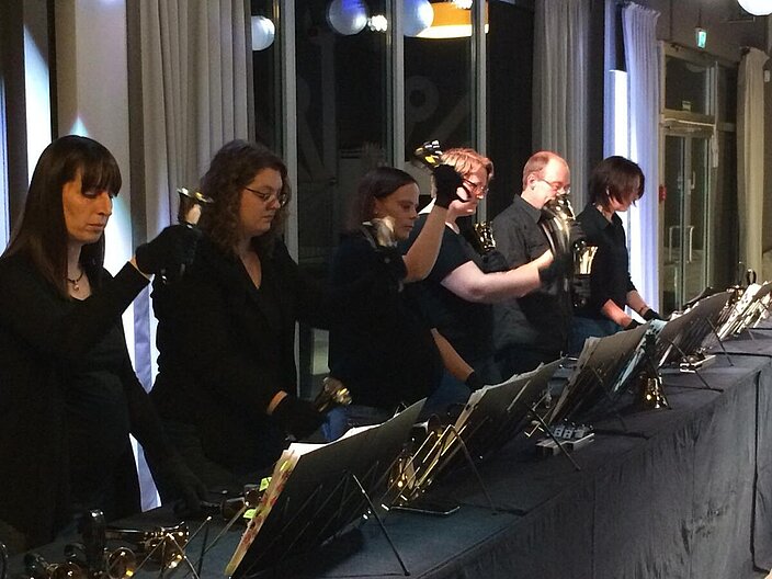 Handglockenchor bestehend aus mehreren schwarz gekleideten Personen mit Handschuhen, die kleine Glocken in den Händen halten.