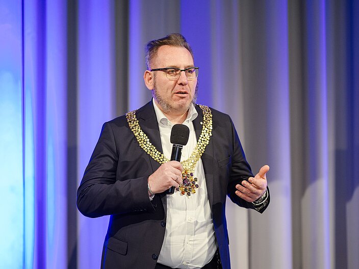 Urkundenverleihung_25_Goldschmiedehandwerk Goldschmied auf der Bühne mit geschmiedeter Kette über die Schultern gelegt.