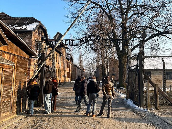 Schüler*innen stehen vor dem Eingang ins Konzentrationslager Auschwitz. Über ihnen steht der Schriftzug "Arbeit macht frei".