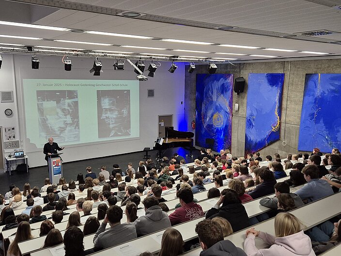 In einem Vorlesungssaal sieht man Klaus Schilling, den Bundeskoordinator der UNESCO-Projektschulen, auf der Bühne vor einer Projektion zu der Rosenburg-Ausstellung sprechen. Der Raum ist voll besucht und alle Tischreihen sind gut mit Schüler*innen esetzt.