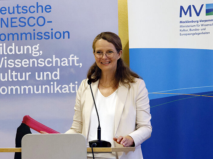 Ministerin Martin steht am Pult. Hinter ihr die Roll-ups von Ministerium und DUK