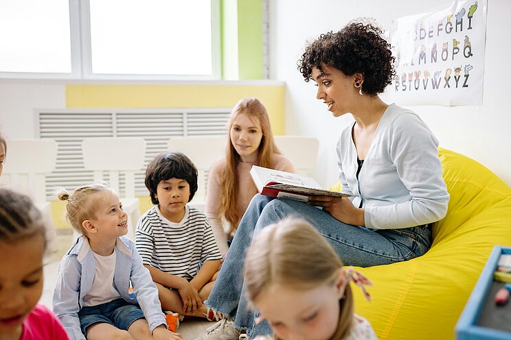 Frühkindliche Bildung_Vorlesen Eine Frau mit schwarzen Locken sitzt auf einem Sessel und liest mehreren Kindern im Kindergarten- oder Grundschulalter vor.