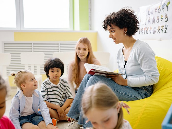 Eine Frau mit schwarzen Locken sitzt auf einem Sessel und liest mehreren Kindern im Kindergarten- oder Grundschulalter vor.
