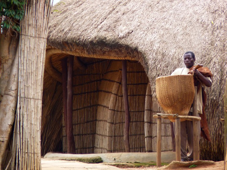 welterbe_uganda_graeber-der-buganda-koenige-in-kasubi_c_CRA-terre Das Bild zeigt ein aus Stroh und Bambus errichtetes Haus vor dem ein Mann mit einem großen geflochtenen Korb steht.