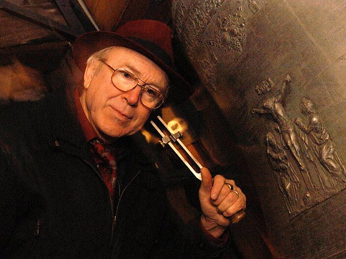Person hält ihr Ohr an die Außenwand einer großen Glocke und prüft den Klang mit einer Stimmgabel.