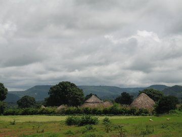 Ein Dorf mit großen, runden Häusern mit schilfartigem, spitzen Dach. Die Landschaft ist sehr grün. Im Hintergrund erkennt man grüne, mittelhohe Berge. 