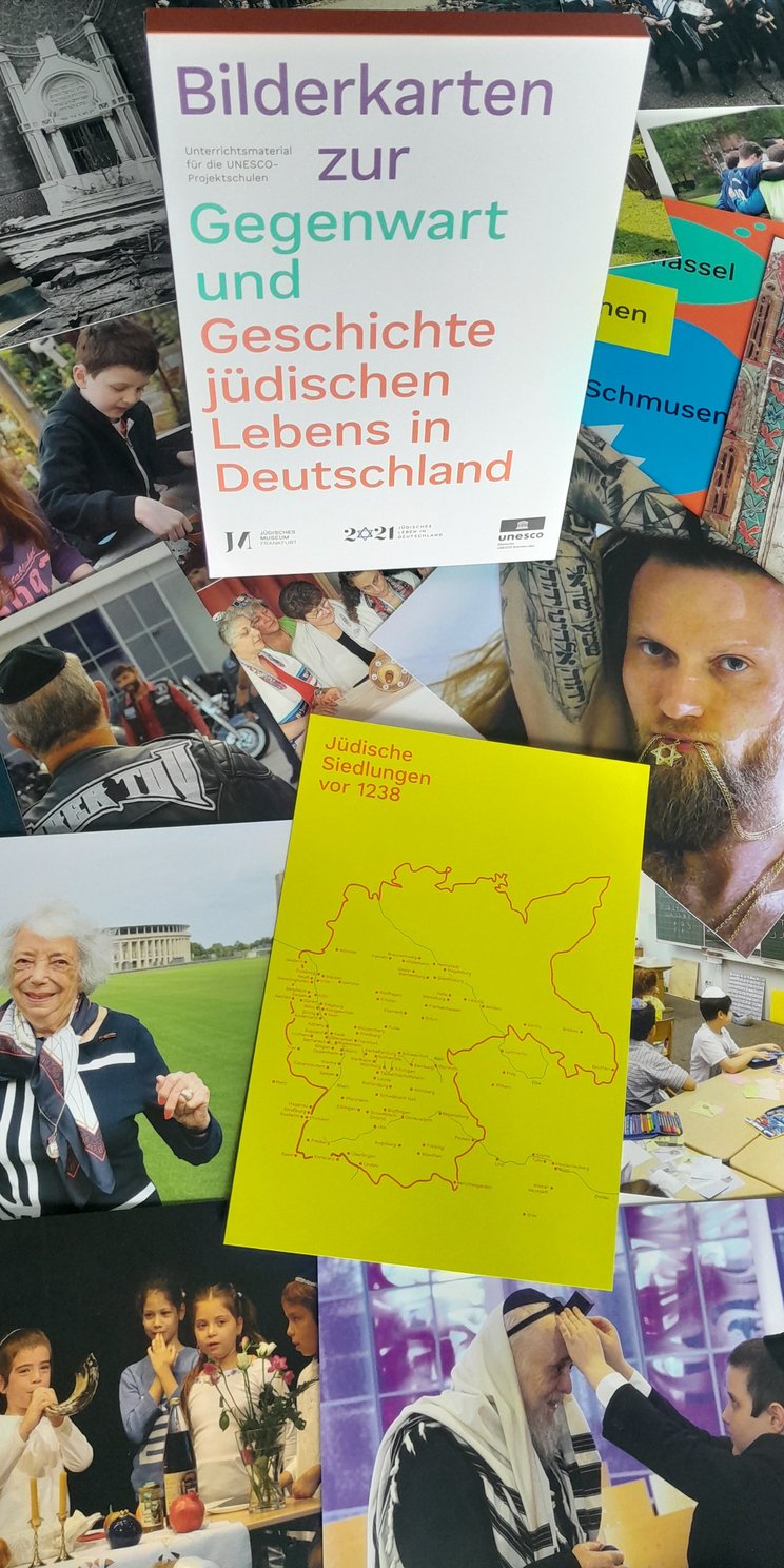 Eine Bilderkarten Box steht in der Mitte und herum liegen viele der einzelnen Bilderkarten mit verschiedenen Fotos zum jüdischen Leben in Deutschland, z.B. das Portrait einer Holocaustüberlebenden, Tattoos in hebräisch, beten etc.