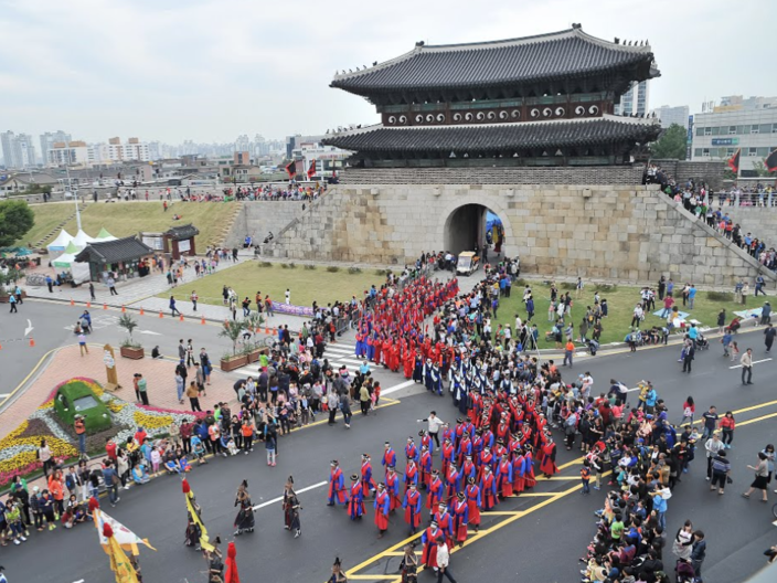From the outside, you can see the entrance to the fortress, which lies behind a road. People in traditional Koranic dress walk festively to the entrance of the fortress. A city can be seen in the background. 