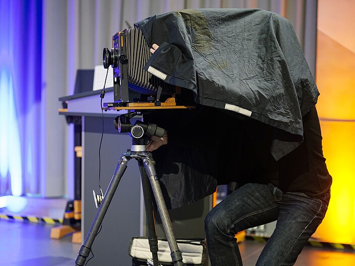 Fotograf steckt unter einer Bedeckung und richtet das Gestell der Analogen Kamera ein.