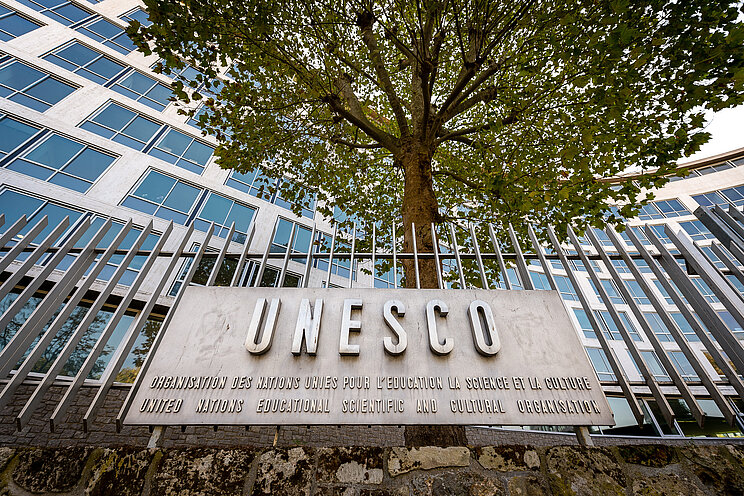 UNESCO Plakette Der Schriftzug UNESCO auf einer bronzenen Plakette, die an einem Stahlzaun befestigt ist. Im Hintergrund ein Baum und eine Gebäudefassade.