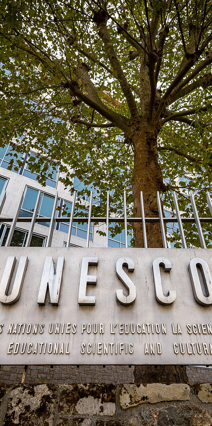 Der Schriftzug UNESCO auf einer bronzenen Plakette, die an einem Stahlzaun befestigt ist. Im Hintergrund ein Baum und eine Gebäudefassade.