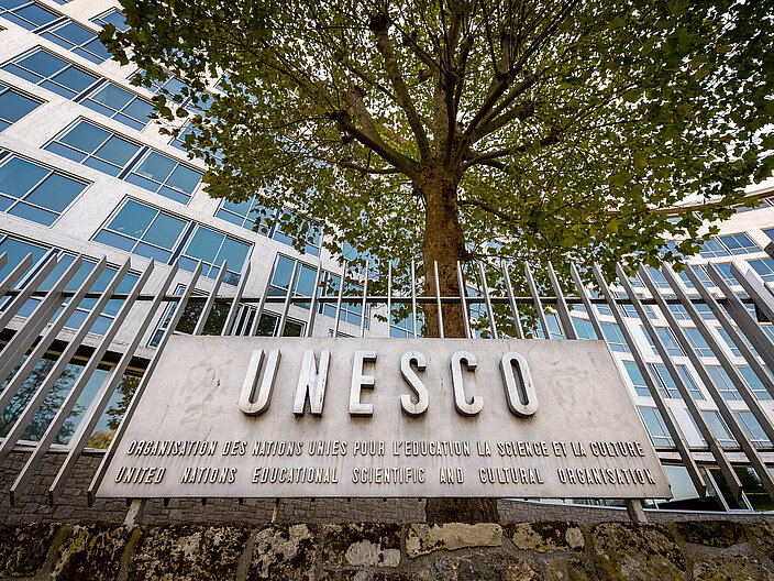 Der Schriftzug UNESCO auf einer bronzenen Plakette, die an einem Stahlzaun befestigt ist. Im Hintergrund ein Baum und eine Gebäudefassade.