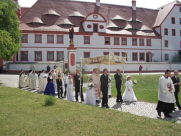 Eine Prozession mit festlich gekleideten Personen zieht &uuml;ber einen gepflasterten Weg. In der Mitte wird ein verzierter Baldachin getragen. Im Hintergrund steht ein gro&szlig;es wei&szlig;-rotes historisches Klostergeb&auml;ude.