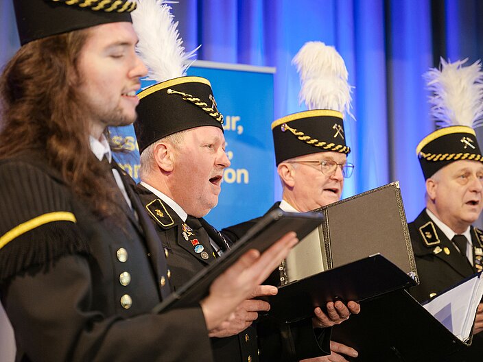 Urkundenverleihung_25_Saarknappenchor Vier Sänger in schwarzer Uniform und Puschel auf dem Kopf singen das Steigerlied.