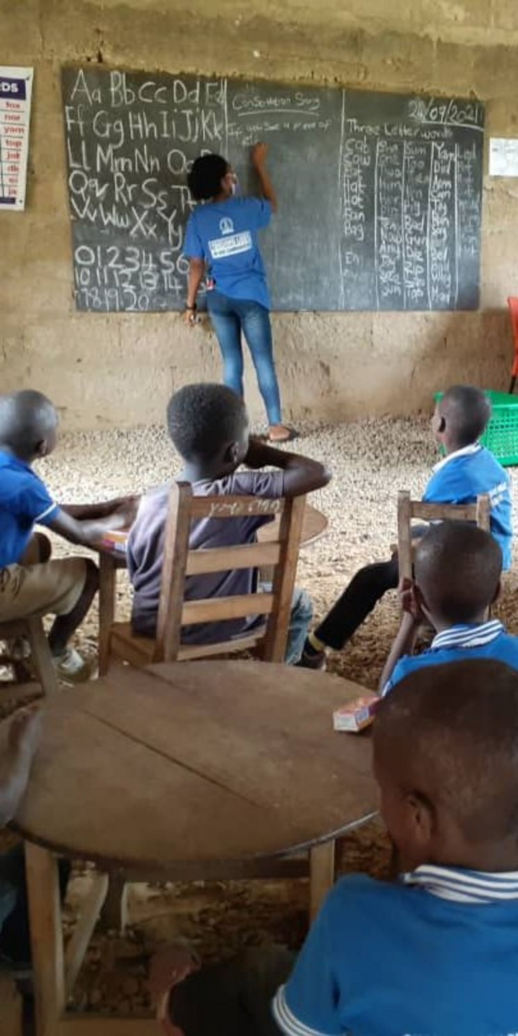 Ein Klassenraum in Ghana. Circa 15 Schülerinnen und Schüler (circa acht Jahre) in blauer Schuluniform sitzen an runden Tischen und schauen in Richtung Tafel. Das Foto ist von hinten aufgenommen. an der Tafel sind eine Projektmitarbeiterin, die Lehrerin und eine weitere erwachsene Person. 