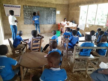 Ein Klassenraum in Ghana. Circa 15 Schülerinnen und Schüler (circa acht Jahre) in blauer Schuluniform sitzen an runden Tischen und schauen in Richtung Tafel. Das Foto ist von hinten aufgenommen. an der Tafel sind eine Projektmitarbeiterin, die Lehrerin und eine weitere erwachsene Person. 