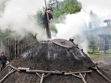Zwei Personen arbeiten an einem gro&szlig;en, rauchenden Kohlenmeiler in einer bewaldeten K&ouml;hlerei. Eine Person steht auf dem Meiler und h&auml;lt eine lange Holzstange, w&auml;hrend dichter wei&szlig;er Rauch aufsteigt.