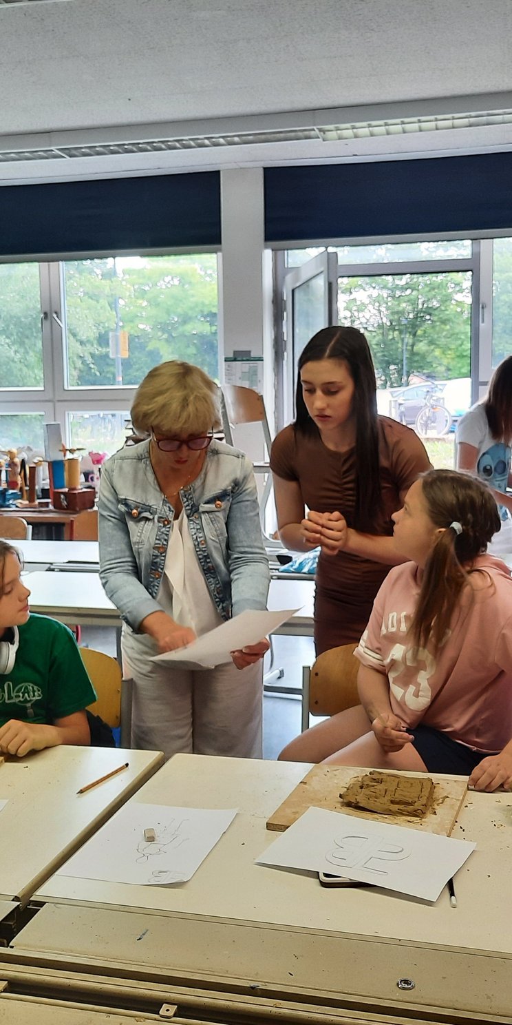 Schülerinnen aus der Ukraine sitzen in einer Klasse und kneten Ton im Rahmen eines Workshops. Eine Lehrerin steht in der Mitte und gibt Erklärungen.