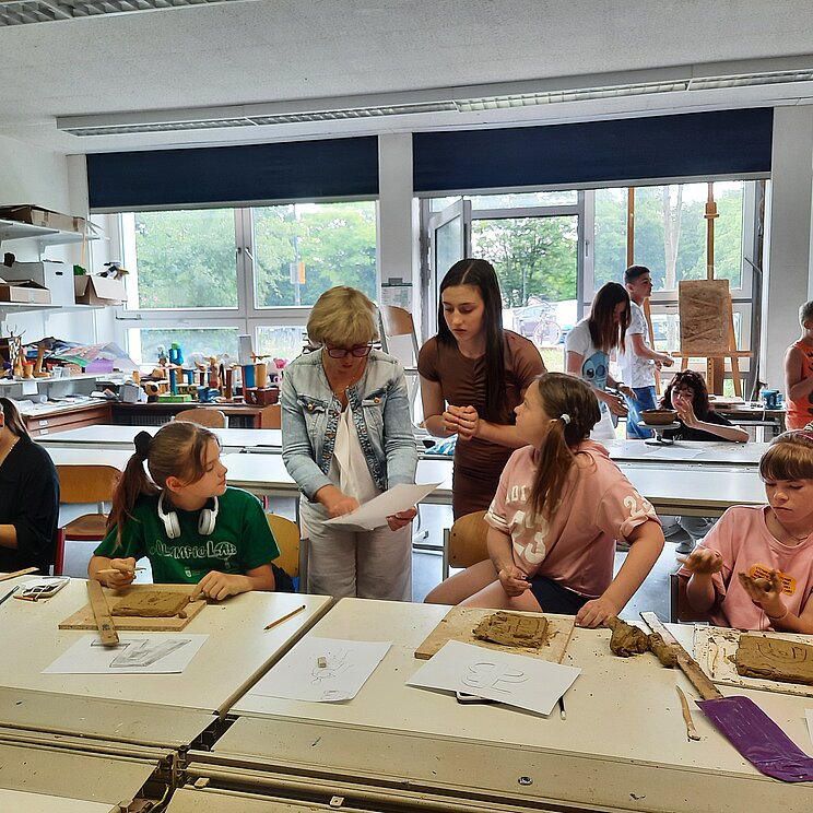 Schülerinnen aus der Ukraine sitzen in einer Klasse und kneten Ton im Rahmen eines Workshops. Eine Lehrerin steht in der Mitte und gibt Erklärungen.