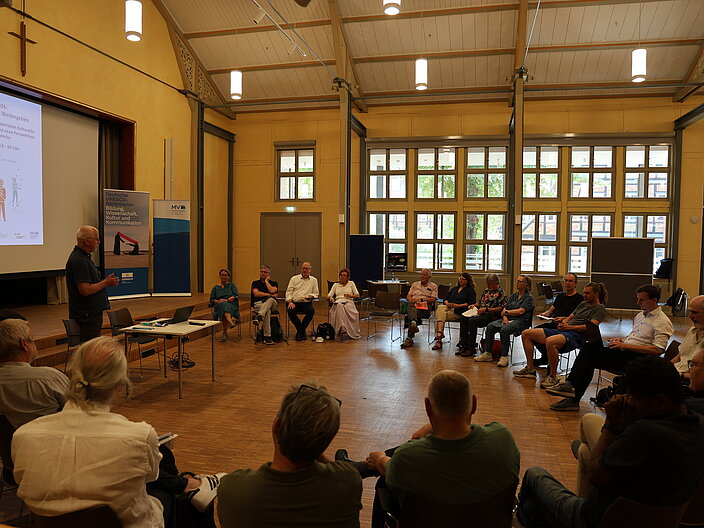 Vortrag aus dem Ministerium. Plenum sitzt in halbkreis um den Vortragenden herum
