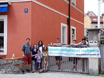 1GENO1_C_Karin_Just_Muenchner_Genossenschaften_web_06 Famile vor Haus mit Banner "spekalutationsfrei wohnen"