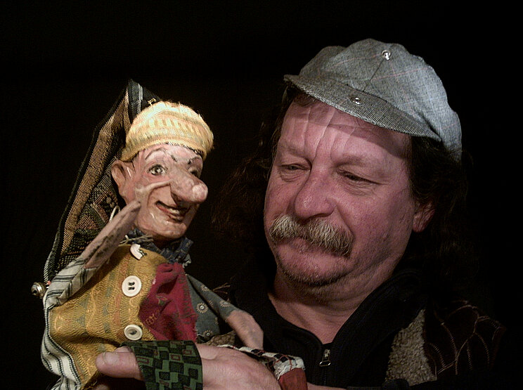 Puppentheater_Höhler Nahaufnahme einer Person mit Schnurrbart und grauer Mütze, die eine handgefertigte Puppe hält. Die Puppe trägt eine Mütze und Kleidung aus verschiedenfarbigen, gemusterten Stoffen. Der Hintergrund ist schwarz.