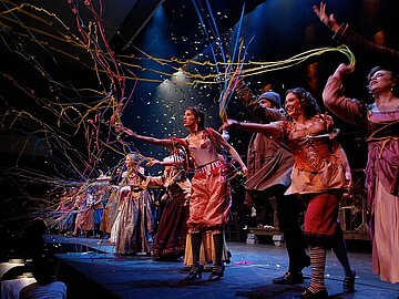 Opernpremiere Das Bild zeigt Schauspieler und Sängerinnen in historischen Kostümen, die sich am vorderen Rand der Bühne in einer Reihe aufgestellt haben. Sie werfen Konfettis und Luftschlangen in den Publikumsraum. Die Personen vorne lachen.
