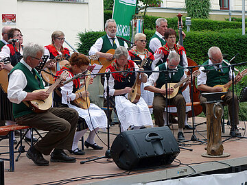 Musikgruppe bei einem Auftritt im Freien. Im Vordergrund spielen mehrere Personen auf Waldzithern, weitere begleiten auf Gitarren und anderen Saiteninstrumenten.
