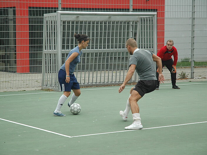 Ausschnitt von einem Bolzplatz. Eine Person verteidigt das Tor. Im Vordergrund versucht eine Frau den Ball an einem Gegenspieler vorbeizuspielen.