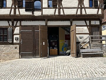 Historisches Fachwerkgeb&auml;ude mit wei&szlig; verputzten Gefachen und dunklen Holzbalken. Die gro&szlig;en Holztore im Erdgeschoss stehen offen und geben den Blick in den Eingangsbereich frei. Vor dem Geb&auml;ude liegt ein gepflasterter Platz, rechts befindet sich eine Holzbank.