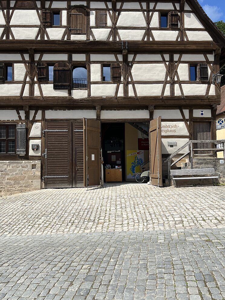 Historisches Fachwerkgeb&auml;ude mit wei&szlig; verputzten Gefachen und dunklen Holzbalken. Die gro&szlig;en Holztore im Erdgeschoss stehen offen und geben den Blick in den Eingangsbereich frei. Vor dem Geb&auml;ude liegt ein gepflasterter Platz, rechts befindet sich eine Holzbank.