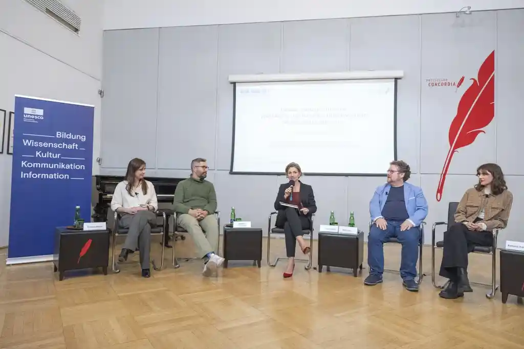 Fünf Menschen sitzen nebeneinander auf Stühlen und diskutieren. Im Hintergrund ist eine Leinwand zu sehen, auf der eine Präsentation projiziert wird.