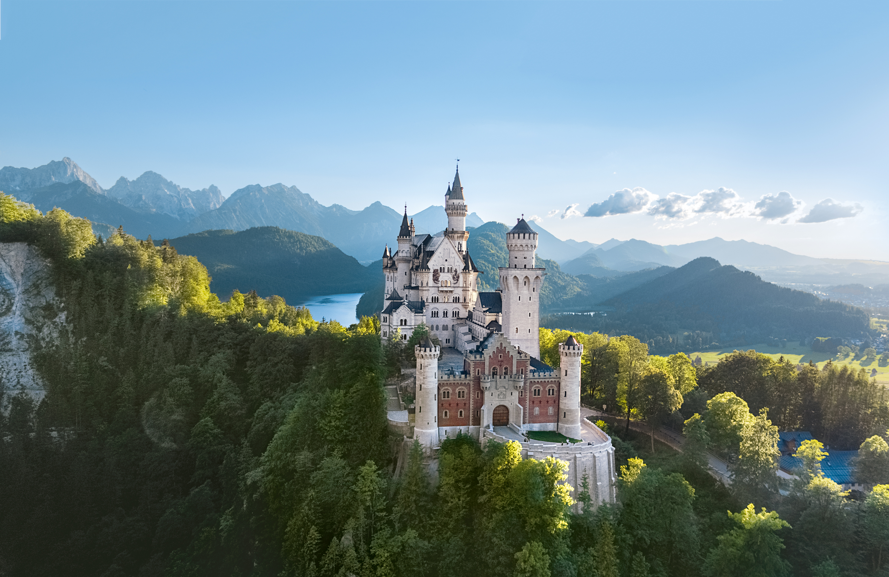 Blick auf das Schloss Neuschwanstein vom Osten aus - auf einem Berg zwischen Bäumen, im Hintergeund ein See und das Alpenpanorama