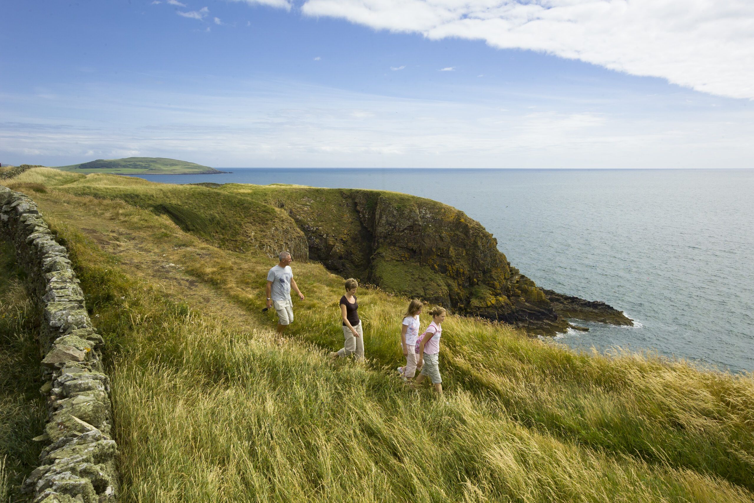 The picture shows two adults and two girls walking along a path in a meadow on the coast. There is a bird's eye view of the people and the sea behind them. 