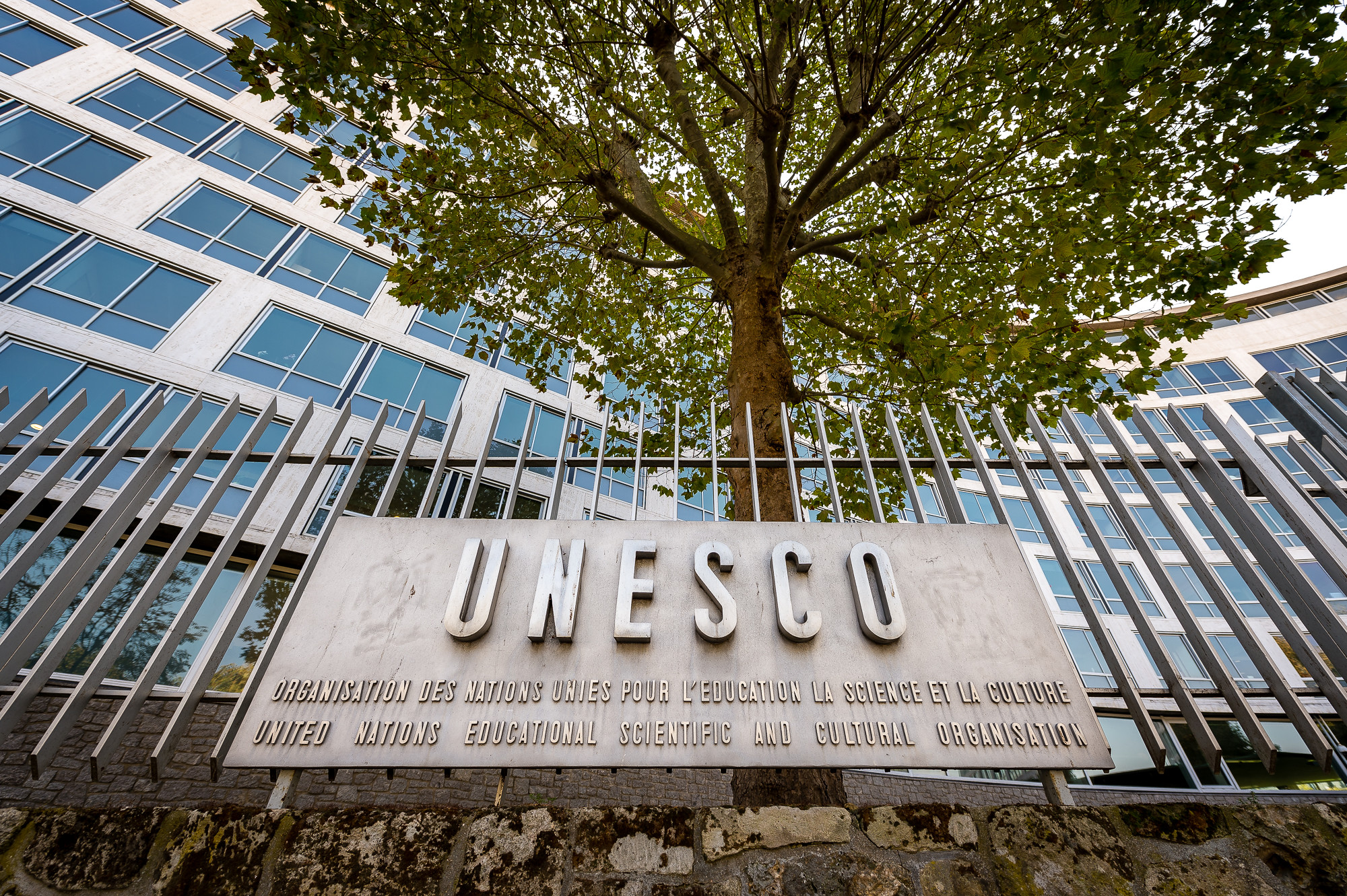UNESCO Plakette Der Schriftzug UNESCO auf einer bronzenen Plakette, die an einem Stahlzaun befestigt ist. Im Hintergrund ein Baum und eine Gebäudefassade.