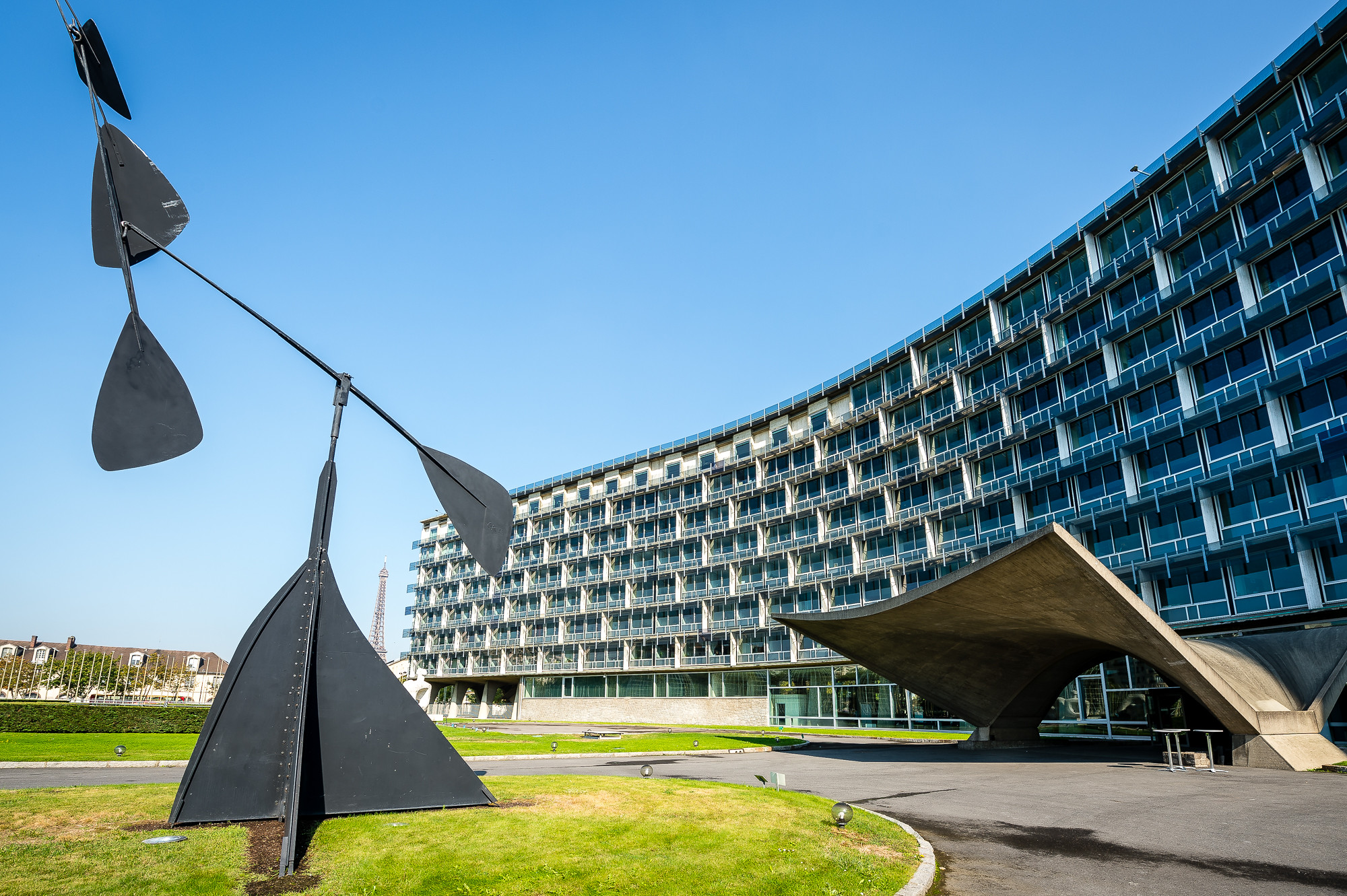 UNESCO Headquarter Blick auf ein konkave Gebäudefront mit expressiven Eingang aus Beton- Auf der linke Bildhälfte eine abstrakte Skulptur aus Stahl.