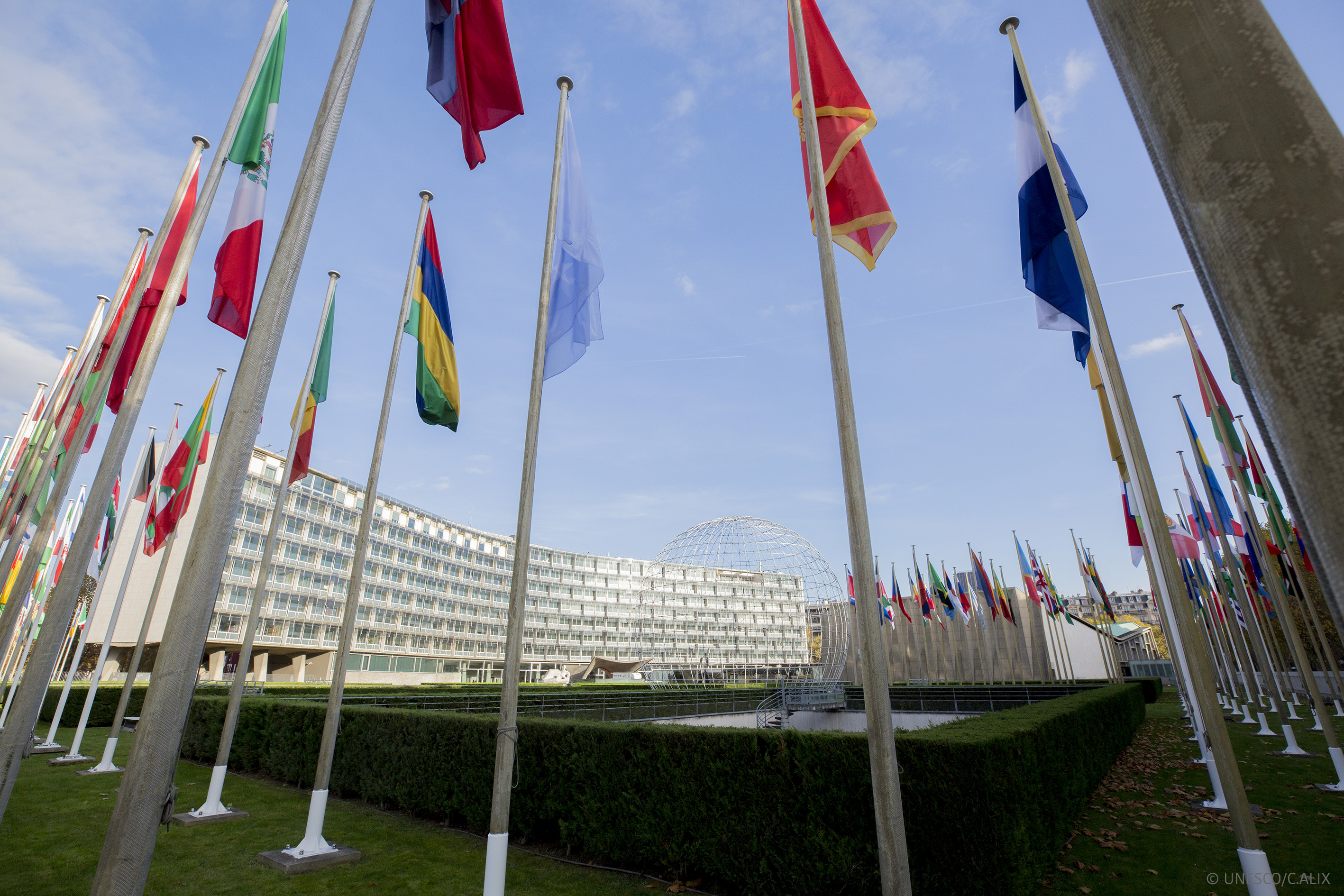 Fahnen am HQ UNESCO Fahnenstangen im Halbkreis, an denen jeweils eine Fahne gehisst ist. Im Hintergrund ein Gebäude.